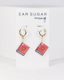 Pink book-shaped earrings with gold hearts on a white card labeled 'Ear Sugar earrings'.