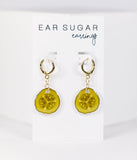 Pair of  pickle slice earrings on a 'Ear Sugar' branded card against a white background